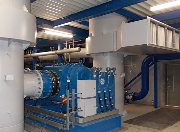 Process air fan installation inside an industrial plant, showing a large blue blower unit connected to white ducts, piping, and support structures in a mechanical room.