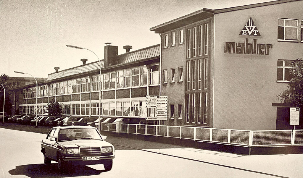 Vintage photo of the MAHLER company building with a long factory facade, the “mahler” sign on the wall, parked cars along the street, and an older sedan in the foreground.
