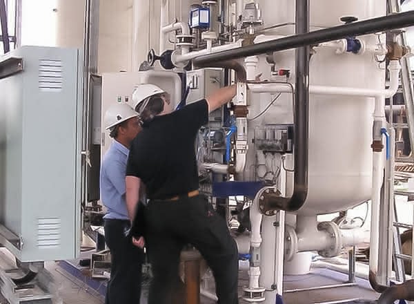 Two technicians in hard hats inspect and adjust a nitrogen generation skid, working beside large white pressure vessels and piping in an industrial plant.