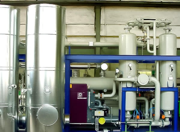 Protective gas system skid with blue steel frame, vertical storage tanks, control cabinet, and interconnected pipes inside an industrial hall.