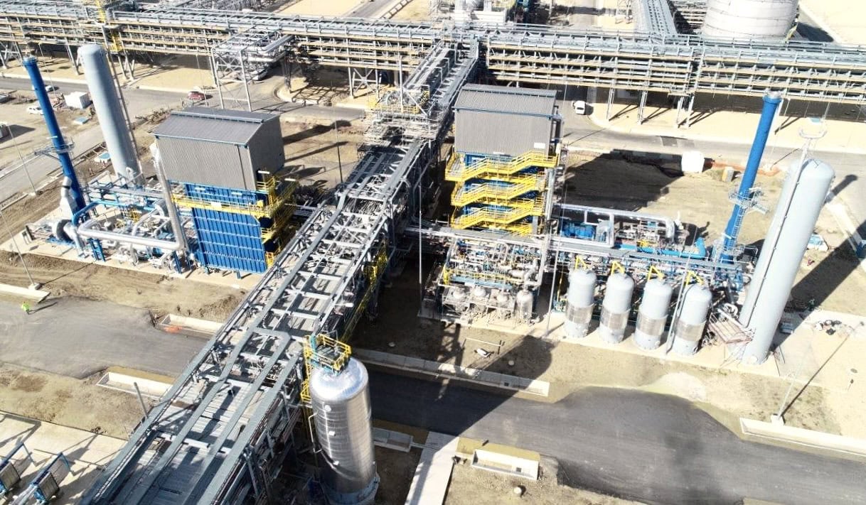 Aerial view of a large industrial processing facility with interconnected pipe racks, tall vertical tanks, blue equipment modules, and yellow stair towers, illustrating a complex engineering and project management site.