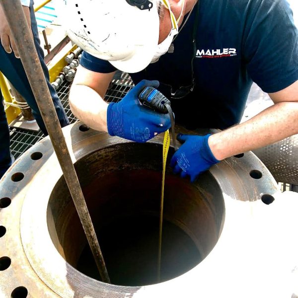 Mahler AGS service technician wearing a hard hat and blue gloves measures the inside of a large metal flange opening on-site, illustrating maintenance and spare parts support.