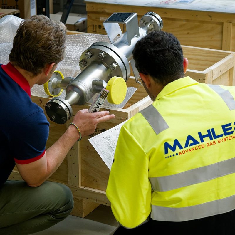 Two MAHLER Advanced Gas Systems employees inspect a large metal component in a wooden crate, reviewing technical drawings in a workshop.