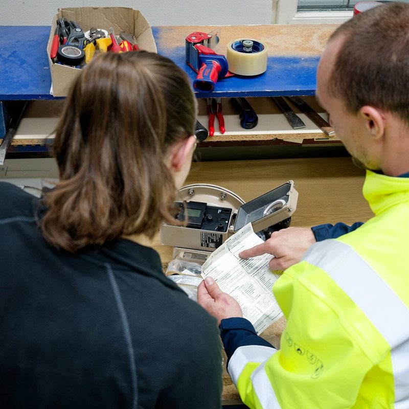 Two MAHLER Advanced Gas Systems employees review documentation together at a workbench while examining a technical device and tools.