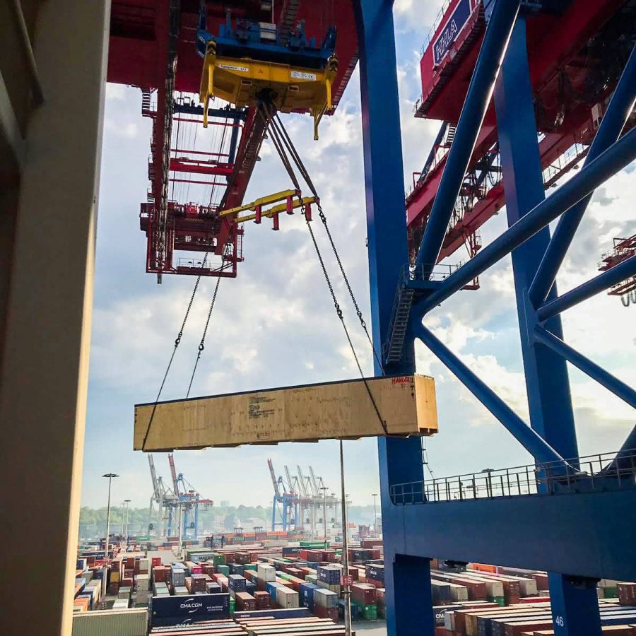 MAHLER equipment crate suspended from a gantry crane above a busy container terminal during shipping.
