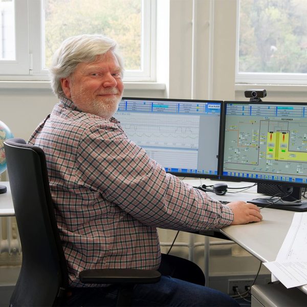 Service engineer at a desk monitors a MAHLER plant remotely, viewing process data and a control system interface on dual computer screens.