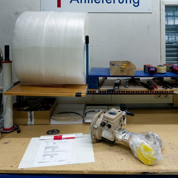 A MAHLER workshop/service bench: on a wooden worktable there are documents and markers, next to a wrapped spare part/assembly (beige metal housing with a flange) and other tools. In the background you can see a large roll of bubble wrap, packing tape, and small parts—looks like service/spare-parts preparation and shipping.