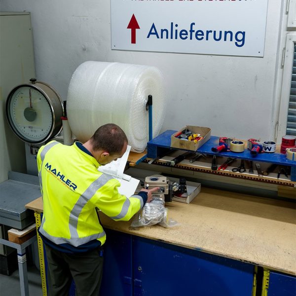 Mahler technician in a high-visibility jacket checks paperwork and inspects a packaged spare part at a workshop bench, with tools and bubble wrap nearby.