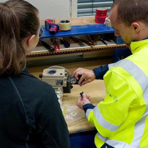 Two MAHLER employees at a workbench examine and assemble a technical spare part, with hand tools and packaging materials visible on the shelf above.