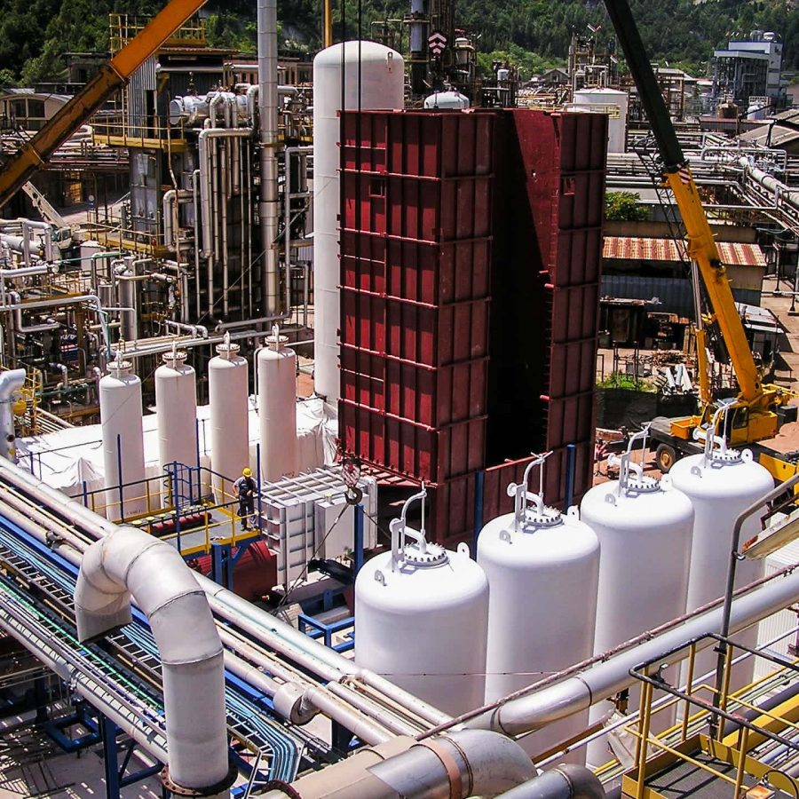 Industrial plant site with cranes lifting a large red module among white storage tanks and dense piping.