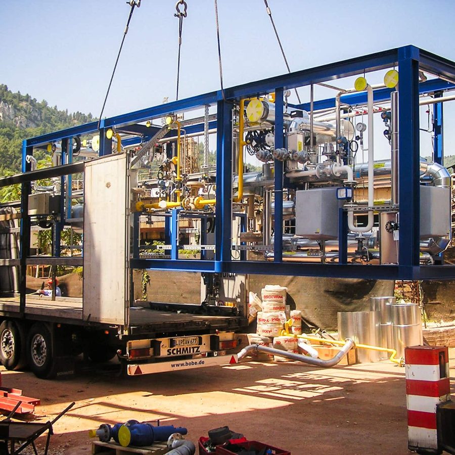 Industrial gas plant module being lifted by crane onto a flatbed truck at an outdoor site.